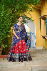Woman in a colorful saree standing outdoors with a yellow building and greenery in the background