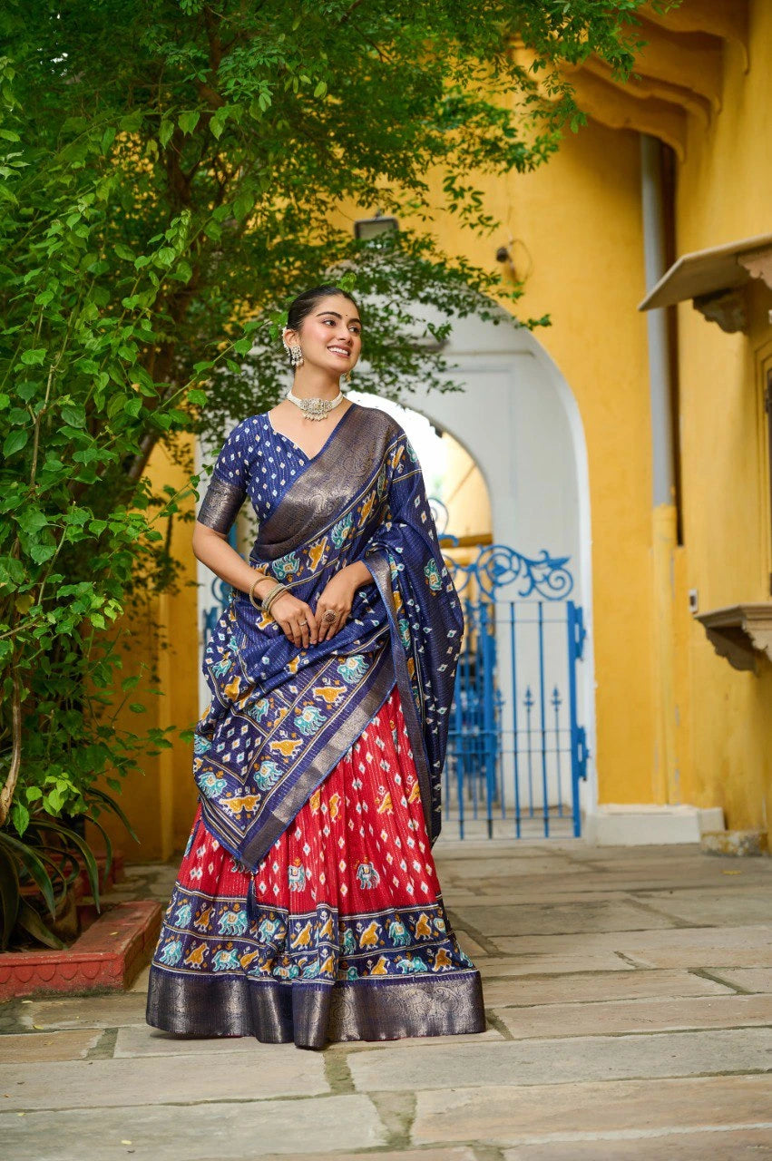 Woman in a colorful saree standing in front of a yellow building with greenery.