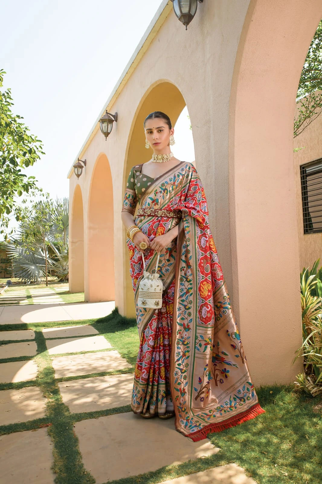 Woman in a colorful saree standing outdoors near a building with greenery.