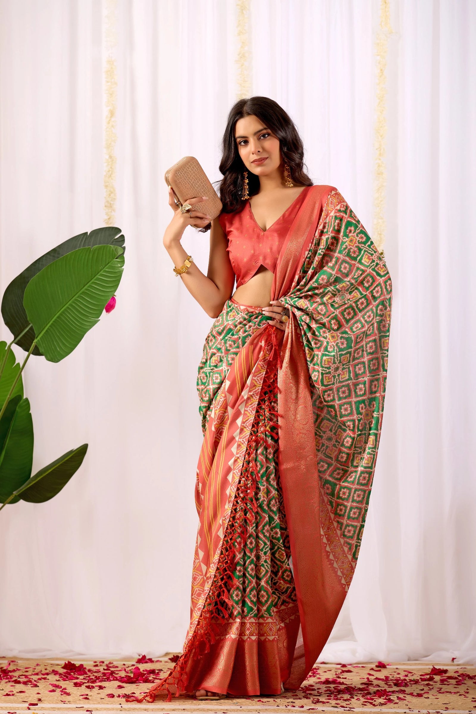 Woman in a red and green patterned saree holding a phone against a white curtain background