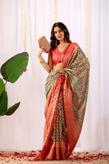 Woman in a red and green patterned saree holding a phone against a white curtain background