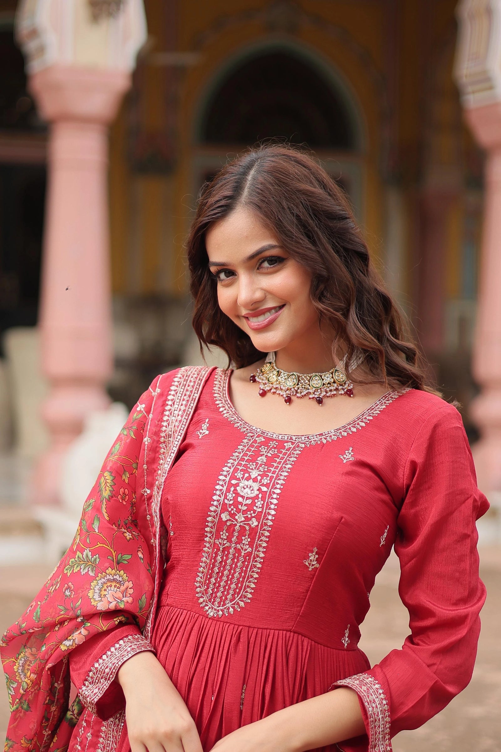 Woman in a red traditional outfit with intricate designs, standing in front of an architectural background.