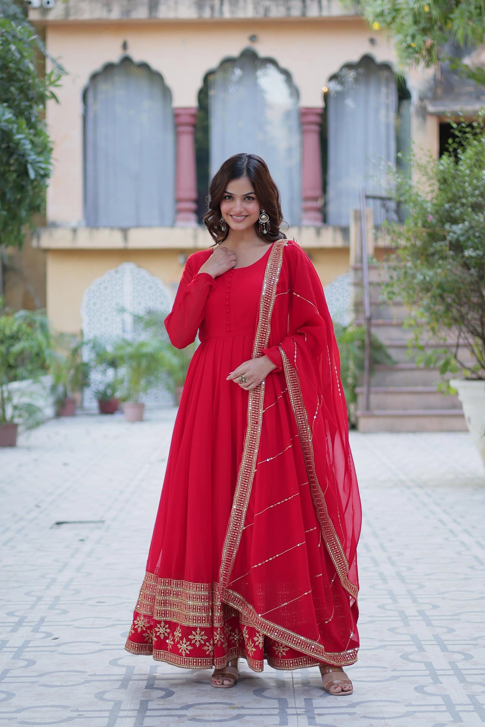 Woman in a red traditional outfit standing outdoors with a building and plants in the background