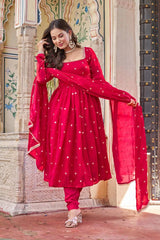 Woman in a red traditional outfit with white patterns standing in front of an ornate architectural background.