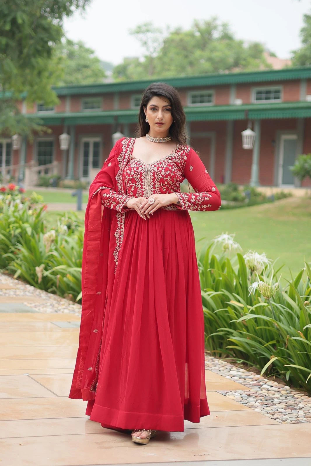 Woman in a red traditional outfit standing outdoors with greenery and a building in the background
