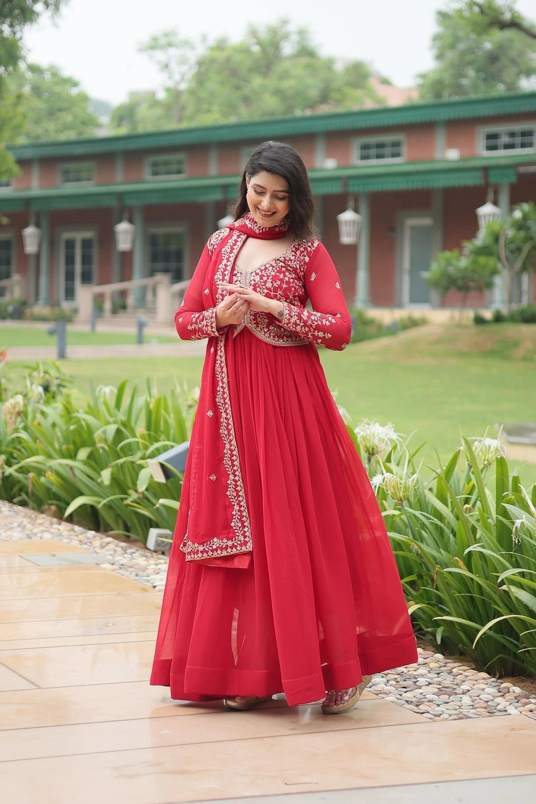 Woman in a red traditional outfit standing outdoors with greenery and a building in the background
