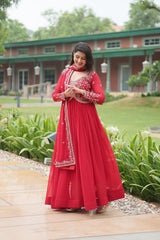 Woman in a red traditional outfit standing outdoors with greenery and a building in the background