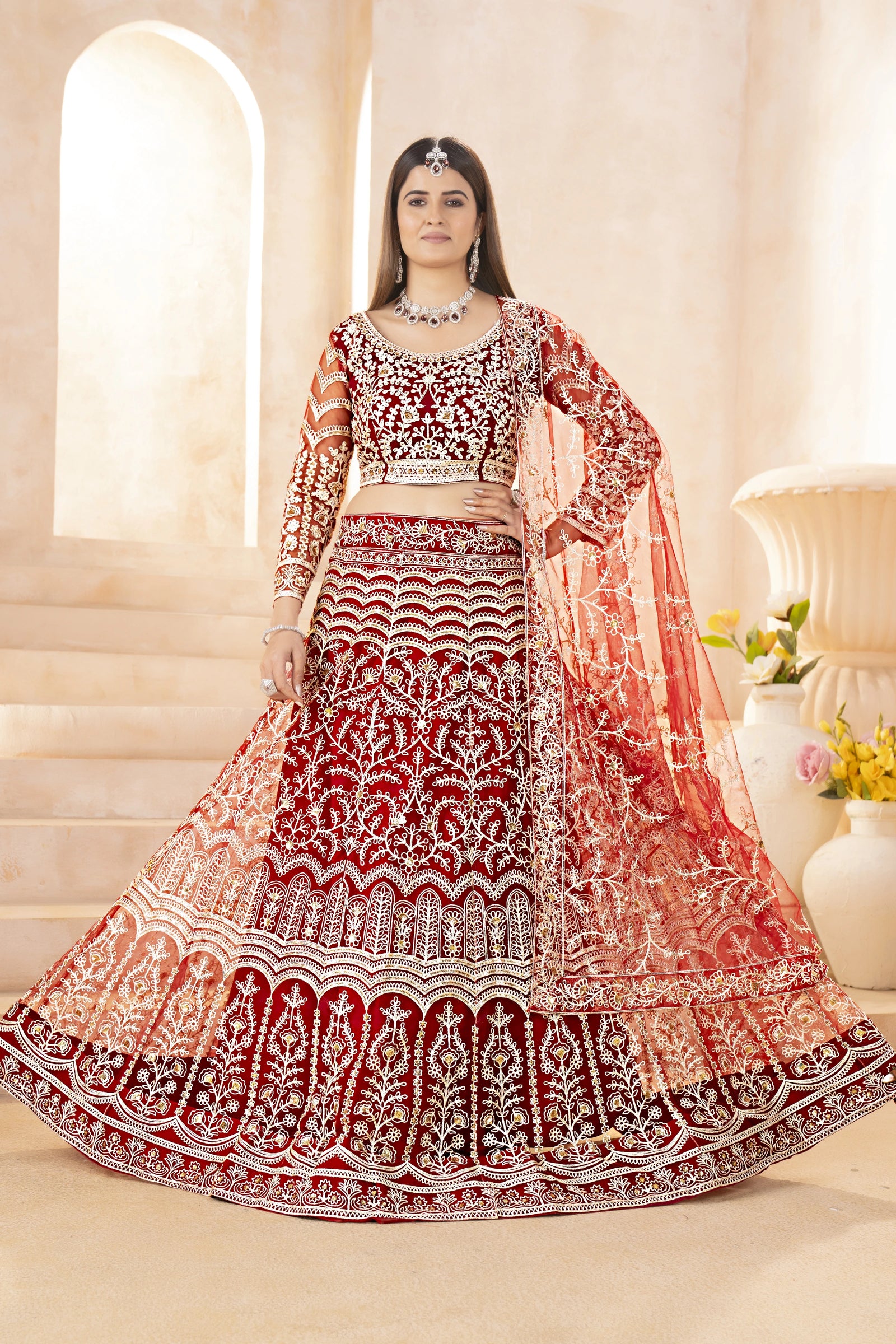 Woman in a red and white embroidered traditional outfit with a matching dupatta, standing indoors.