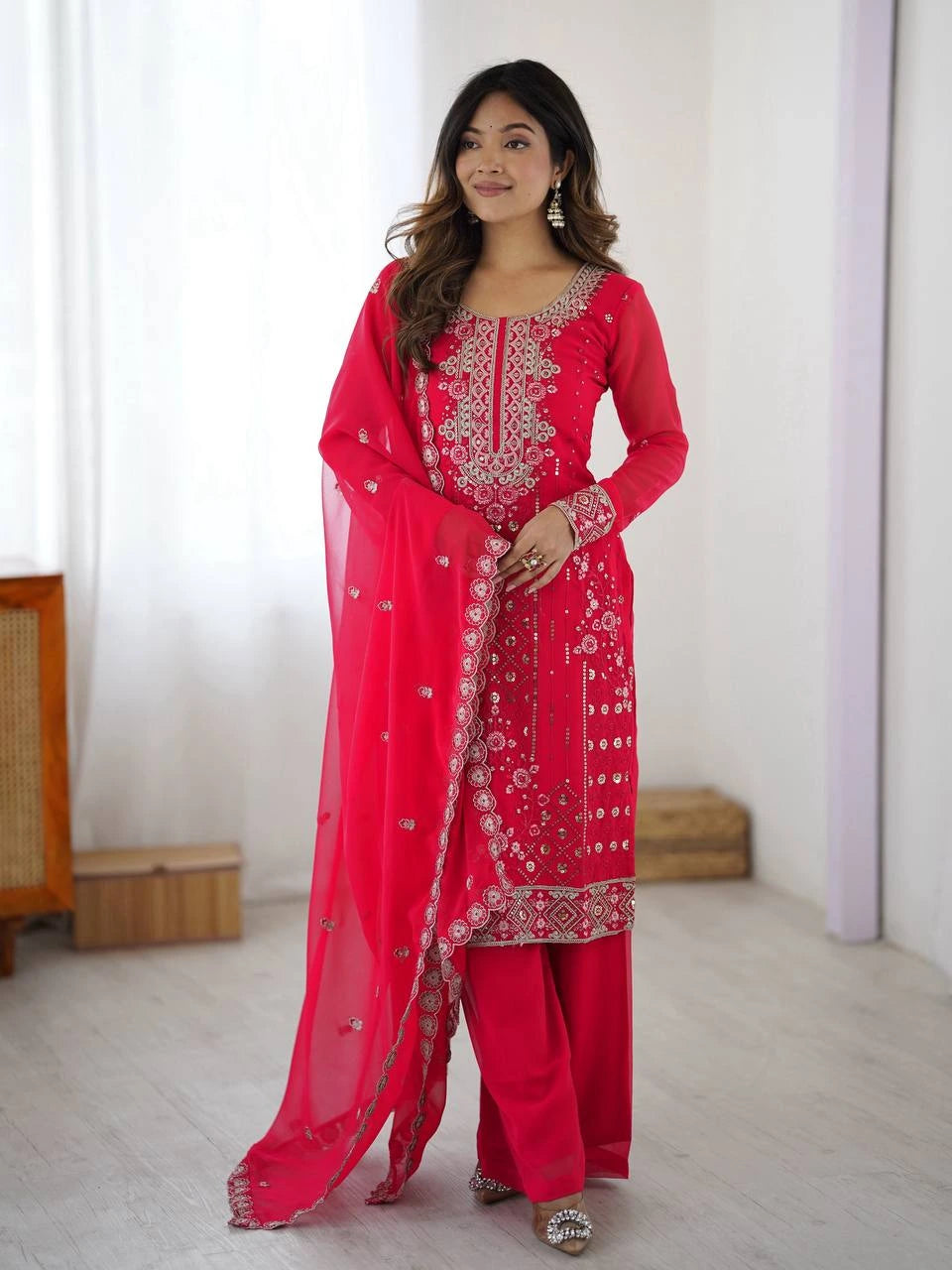 Woman wearing a red traditional outfit with intricate embroidery in a minimalistic room.