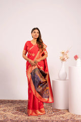 Woman in a red saree with gold border standing in a room with a white wall and decorative vase.