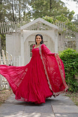 Woman in a bright pink traditional outfit standing outdoors with greenery and a white structure in the background.