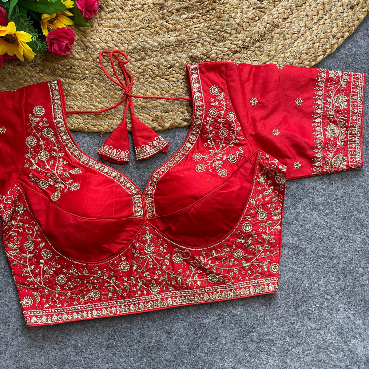 Red heavy silk blouse with intricate gold zari embroidery, deep neckline, short sleeves, and dori tie-up back, displayed on a textured mat with floral accents.