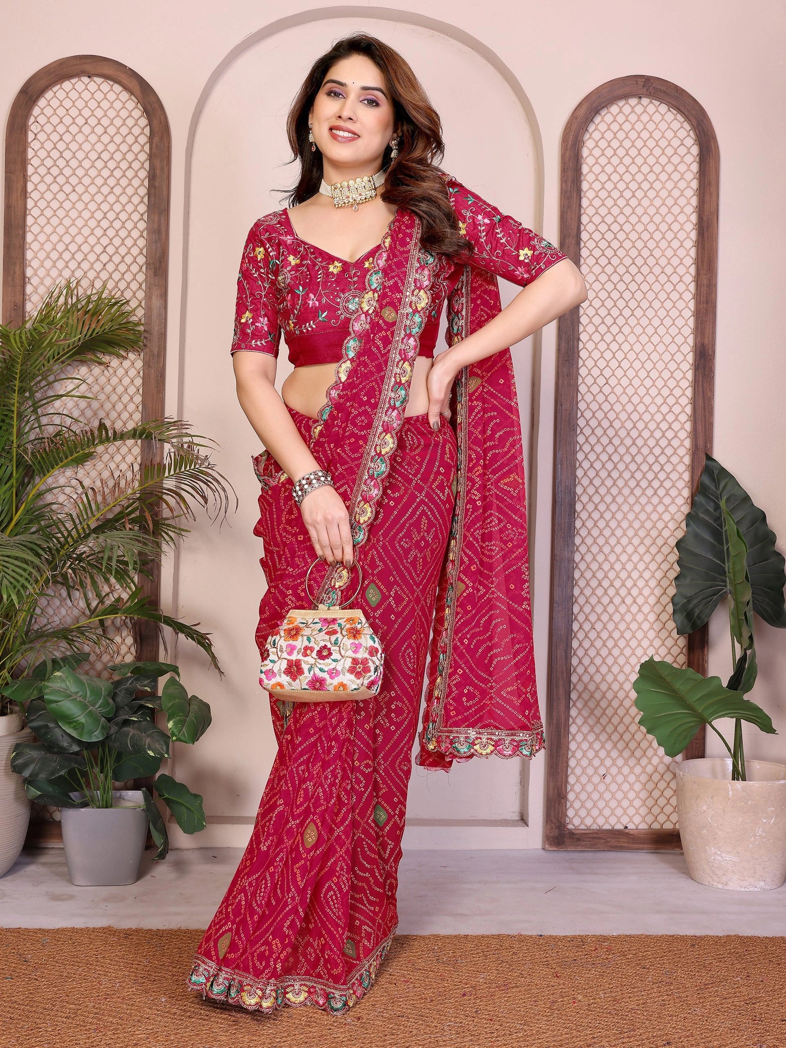 Woman in a red saree with floral patterns standing indoors with plants in the background