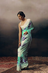 Woman wearing a light green saree with floral patterns on a textured floor.