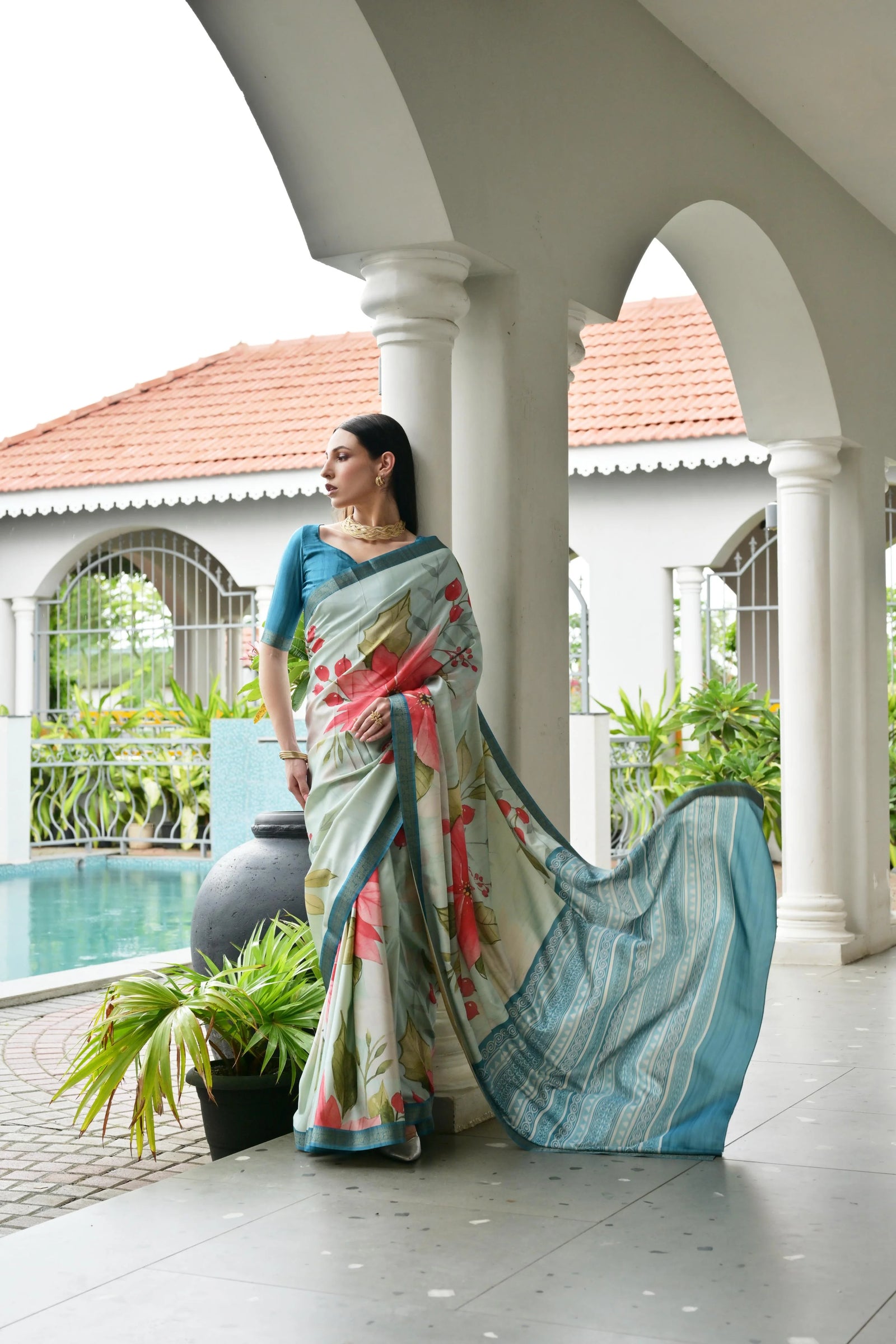 Woman in a floral saree standing on a porch with a pool and garden in the background