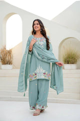Woman in a light blue traditional outfit with floral embroidery standing in a decorative outdoor setting.
