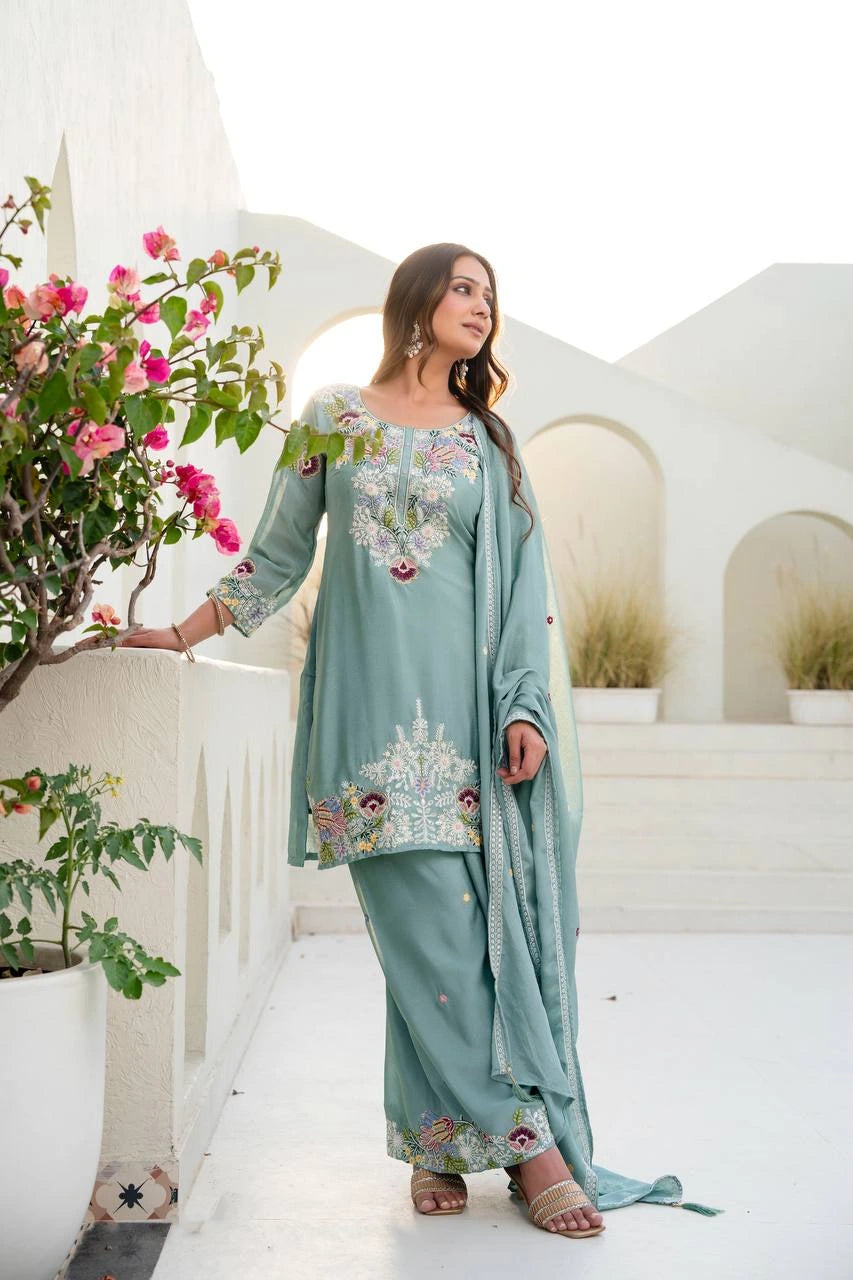 Woman in a light blue embroidered traditional outfit standing in a decorative outdoor setting with flowers and arches.
