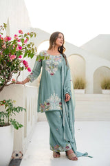 Woman in a light blue embroidered traditional outfit standing in a decorative outdoor setting with flowers and arches.