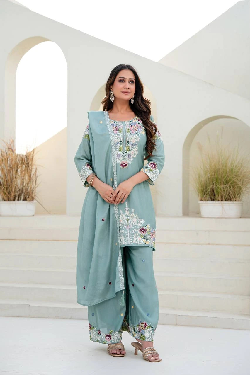 Woman wearing a light blue embroidered traditional outfit standing in a decorative setting.