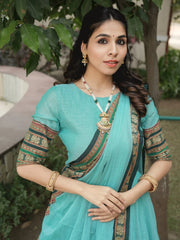 Woman in a turquoise saree with gold jewelry standing outdoors.
