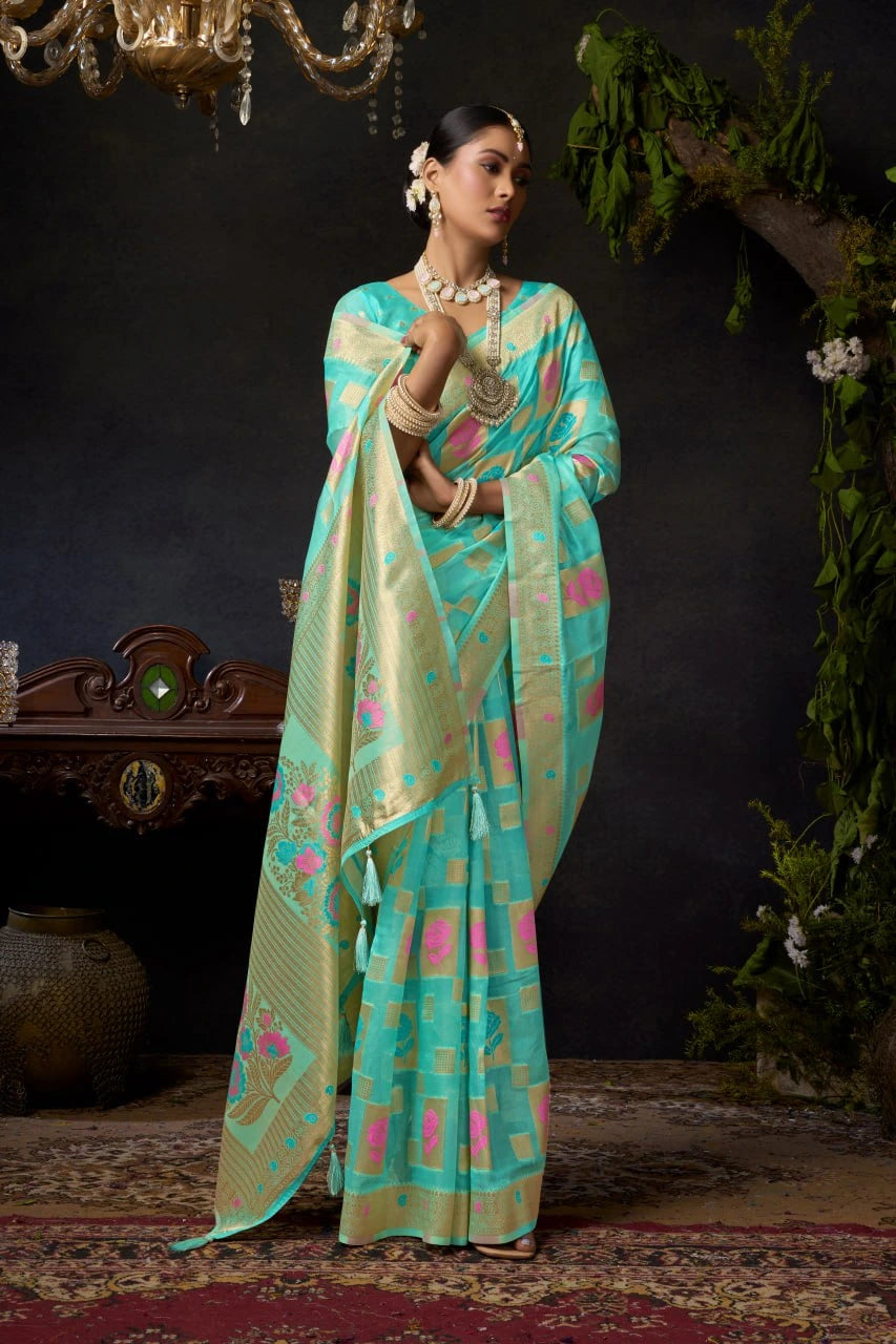 Woman in a turquoise saree with pink patterns standing against a dark background