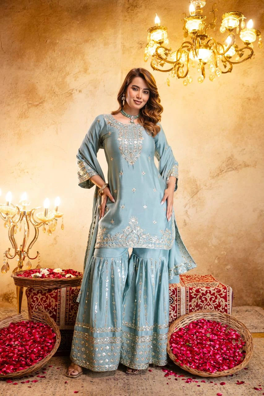 Woman in a light blue traditional outfit standing in a decorated room with chandeliers and flower arrangements.