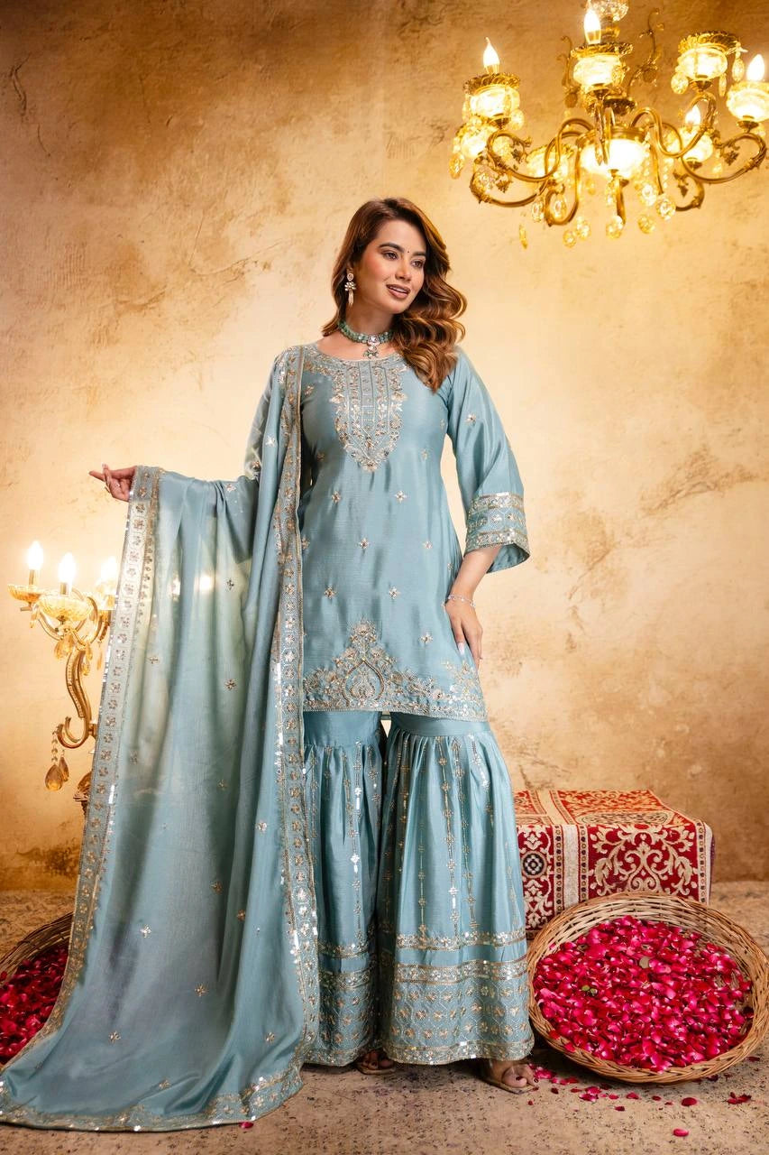 Woman in a light blue traditional outfit with embroidery, standing in a decorated room with chandeliers and floral arrangements.