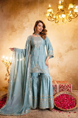 Woman in a light blue traditional outfit with embroidery, standing in a decorated room with chandeliers and floral arrangements.