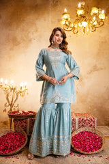 Woman in a light blue embroidered traditional outfit standing in a decorated room with chandeliers and flower arrangements.