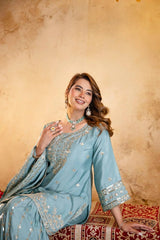 Woman in a light blue embroidered traditional outfit sitting on a patterned rug against a beige wall.