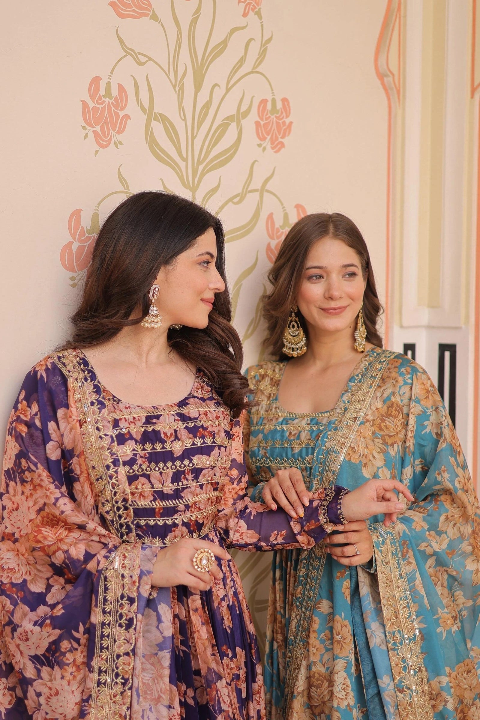 Two women in traditional embroidered dresses standing against a decorative wall.