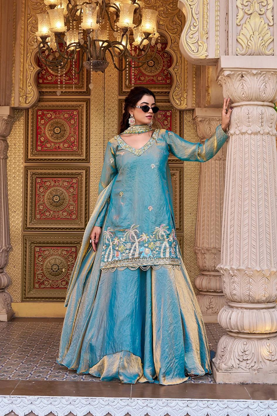 A woman poses elegantly in a sky-blue traditional ethnic outfit inside a royal palace setting.