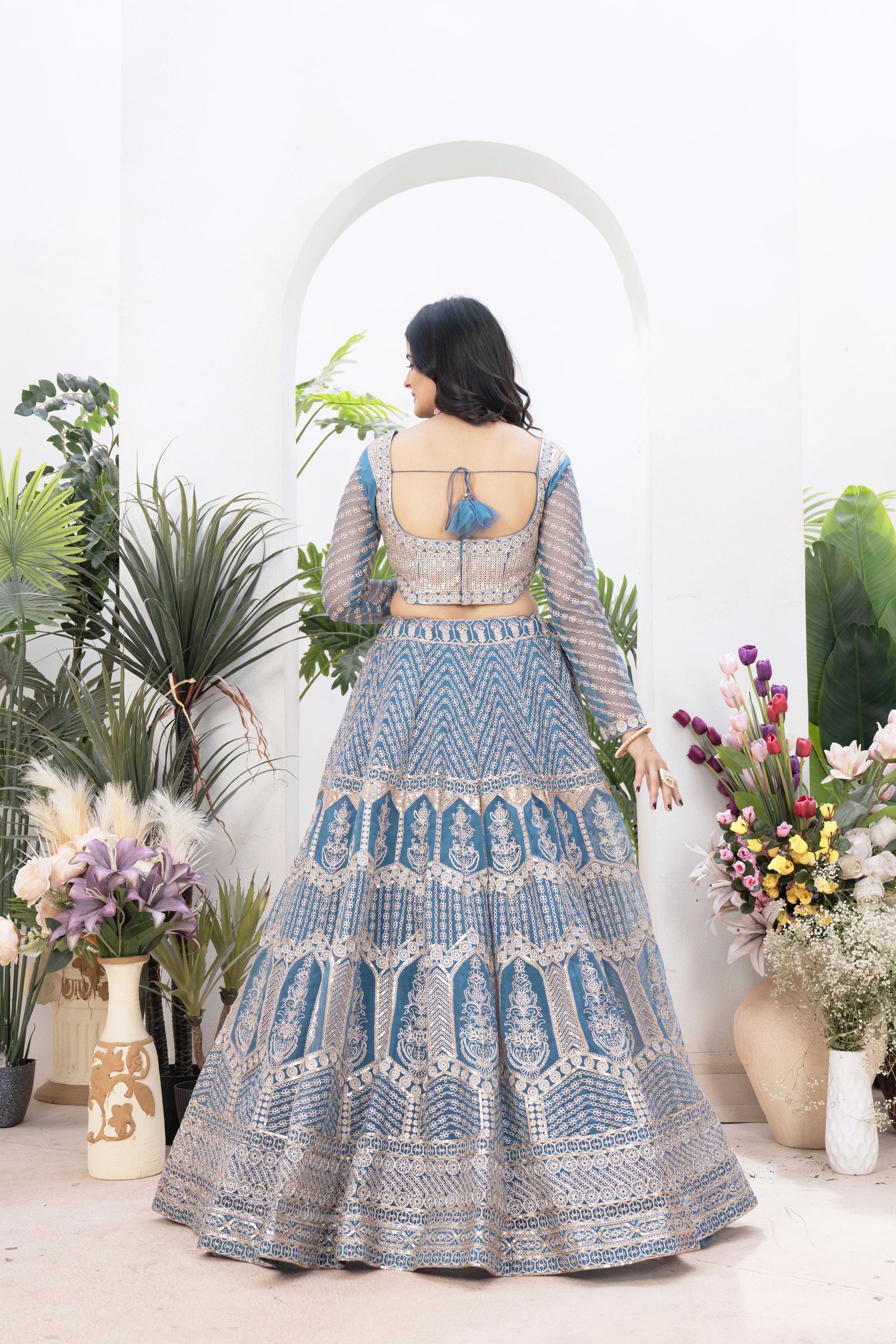 Woman wearing a blue embroidered traditional outfit with a white background and decorative plants.