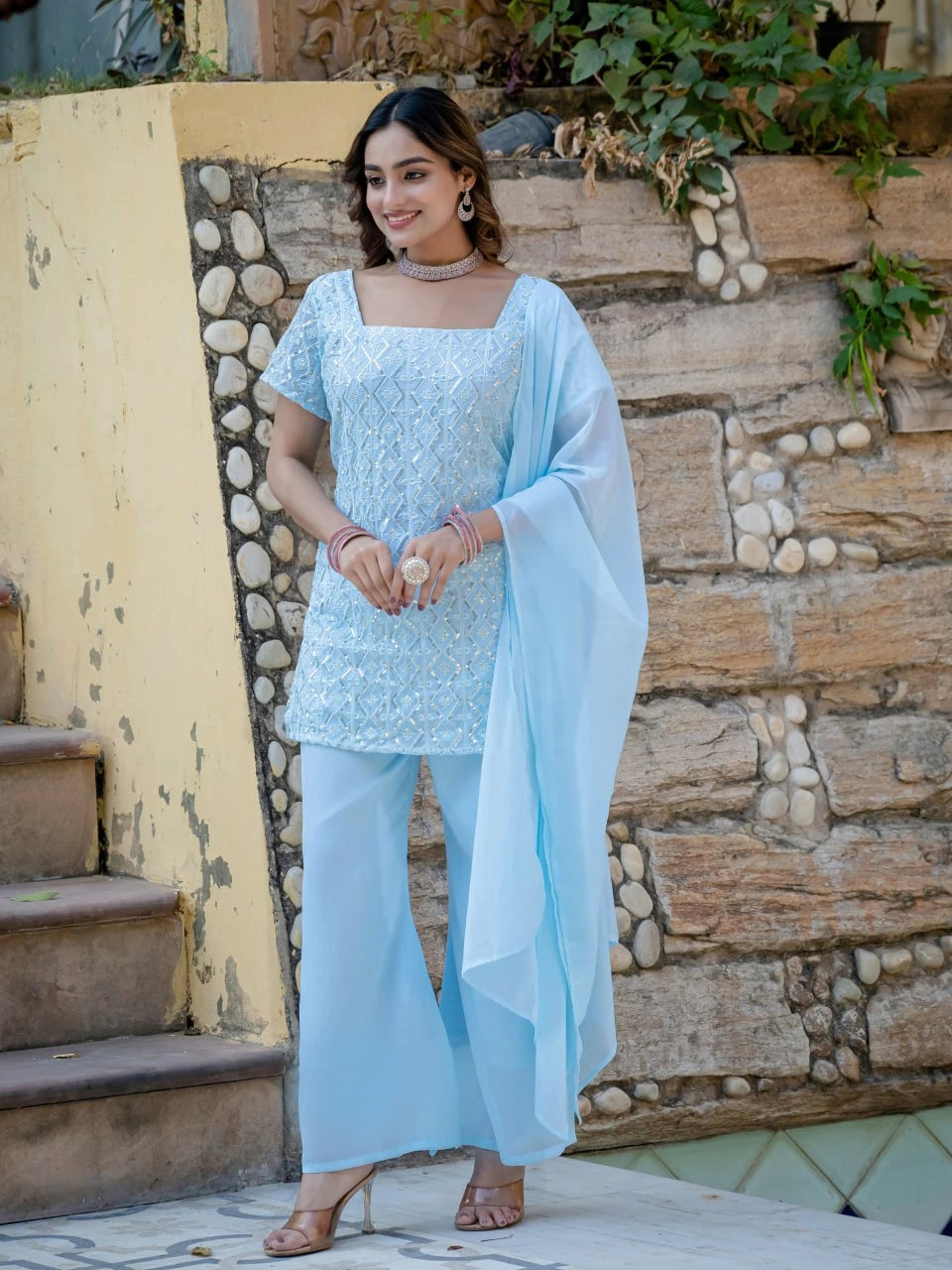 Woman in a light blue traditional outfit standing outdoors against a stone wall.