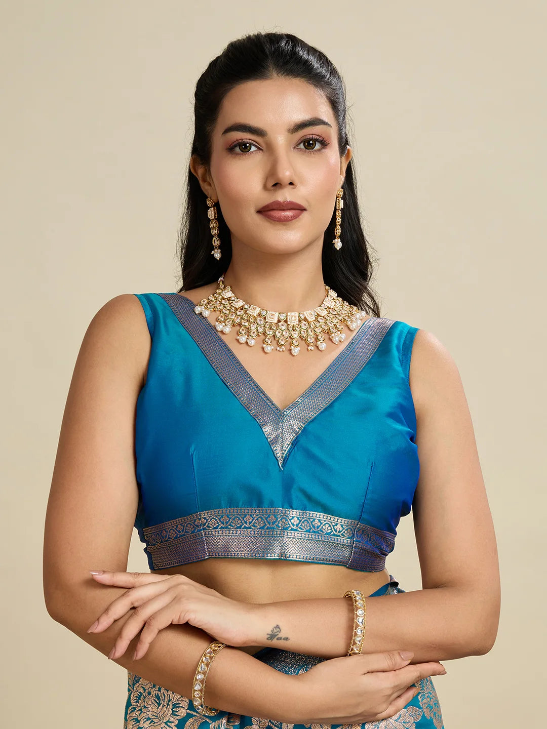 Woman wearing a blue saree with silver border and gold jewelry on a beige background