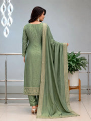 Woman in a green traditional outfit with a matching dupatta in a room with a plant and decorative wall.