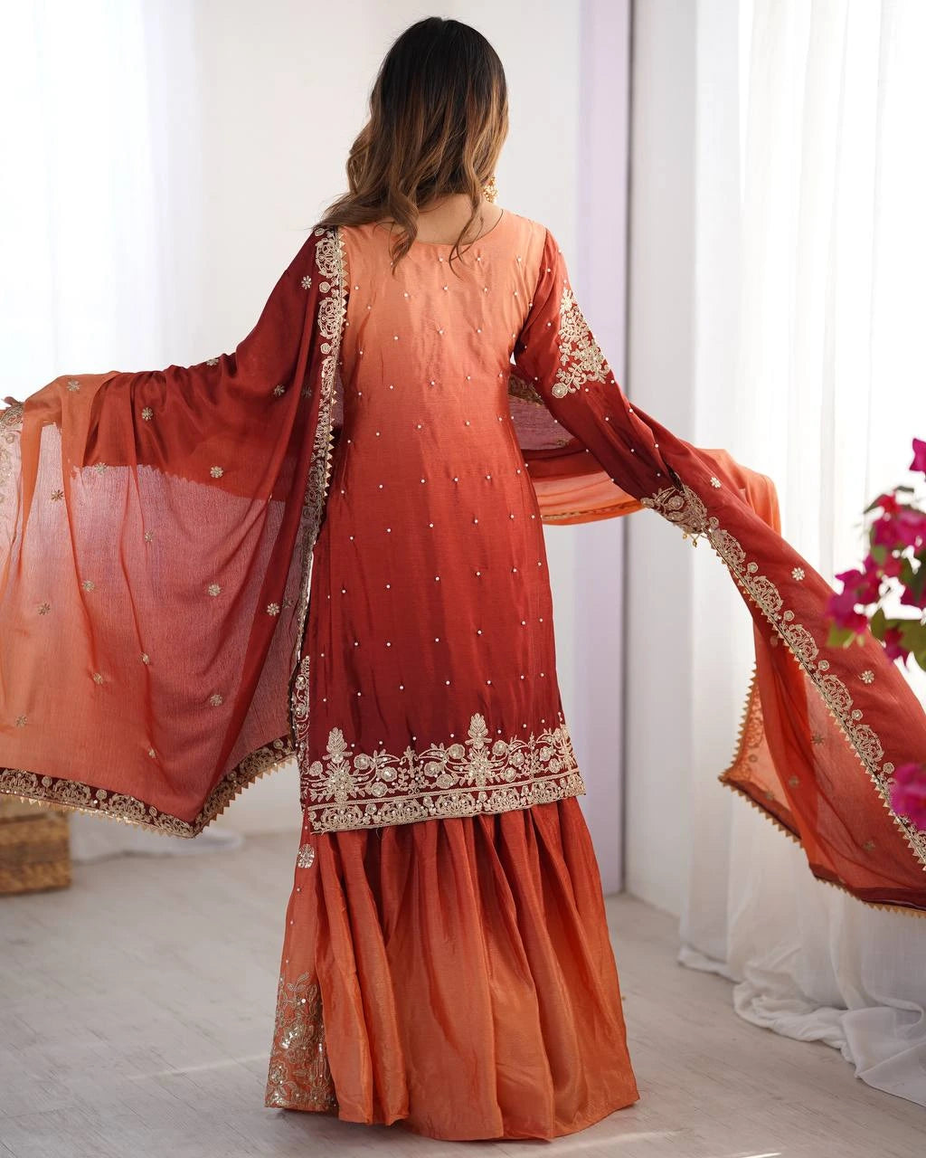 Woman wearing an orange traditional outfit with a sheer dupatta in a minimal indoor setting.