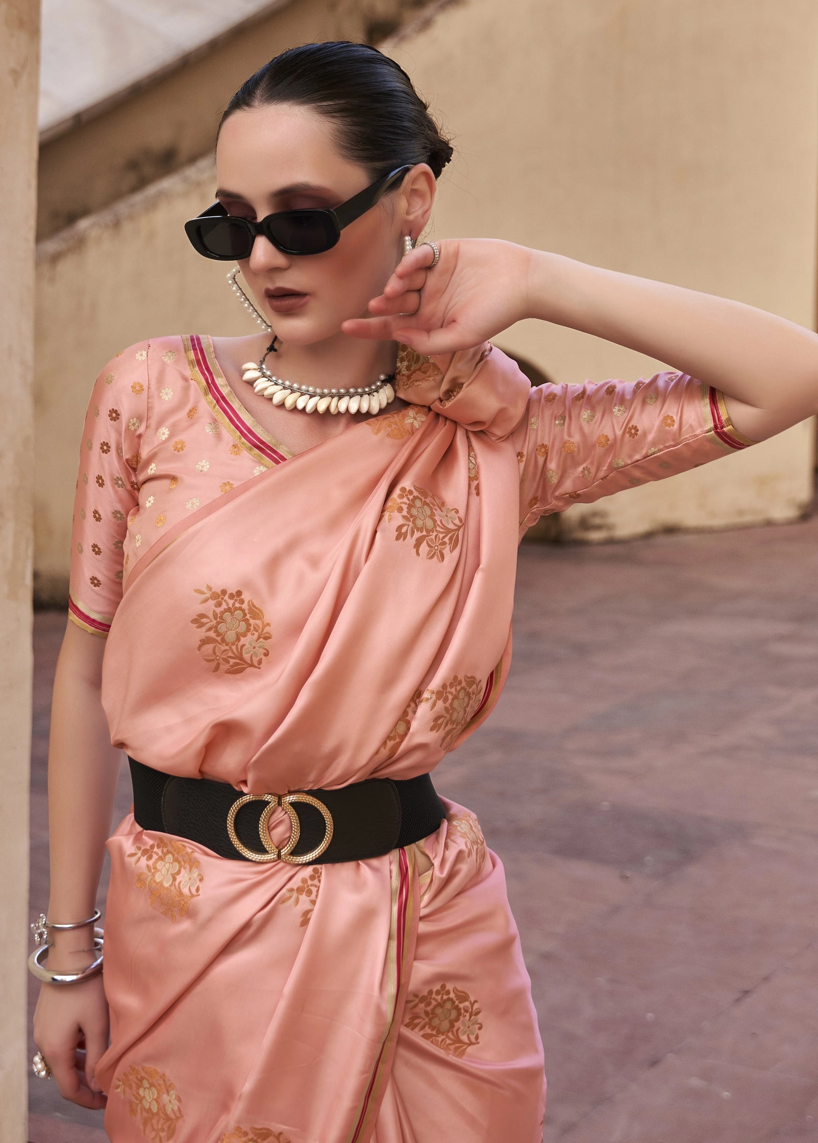 Woman wearing a pink saree with gold patterns, black sunglasses, and a pearl necklace.