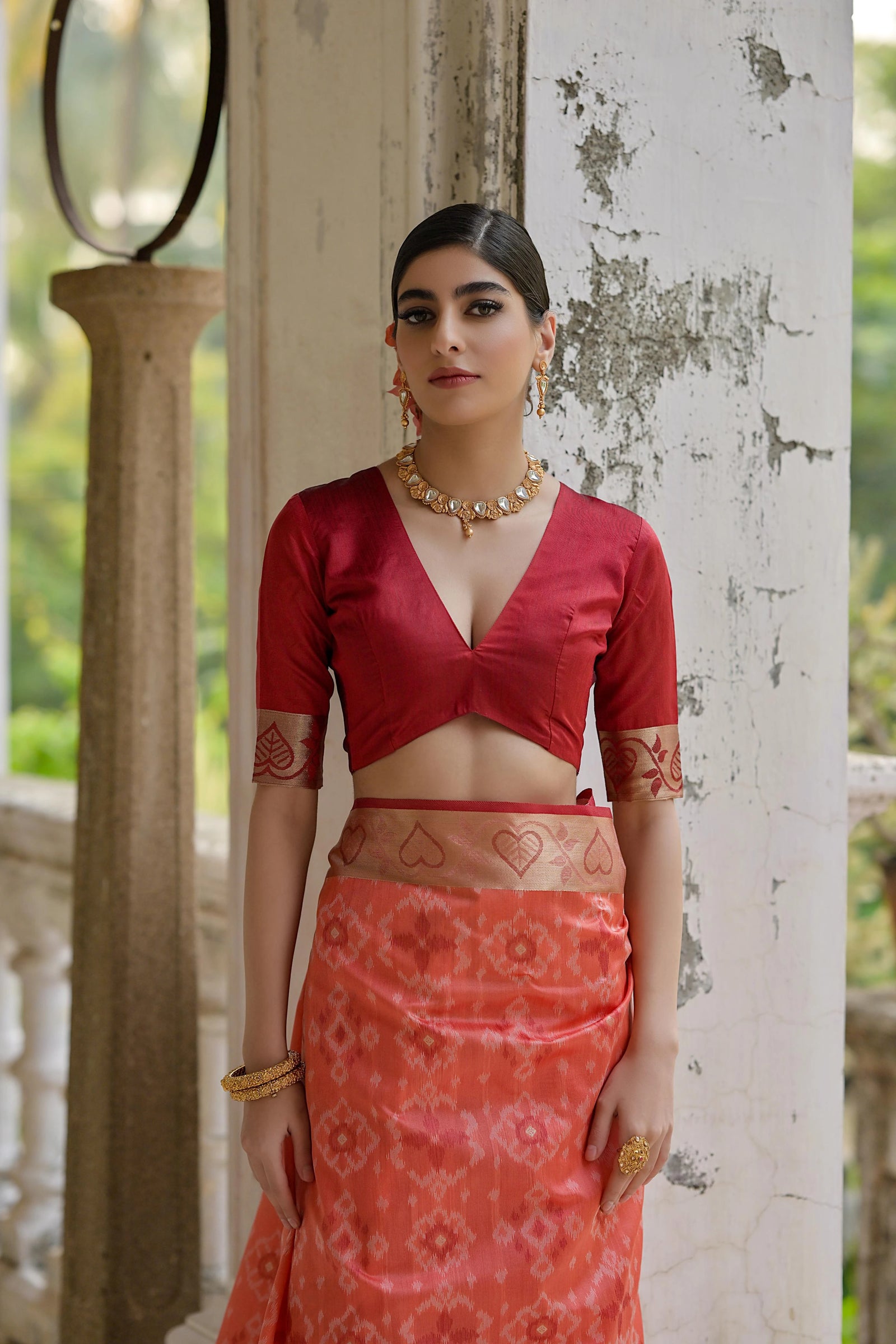 Woman in a red top and patterned skirt standing against a white wall with greenery in the background