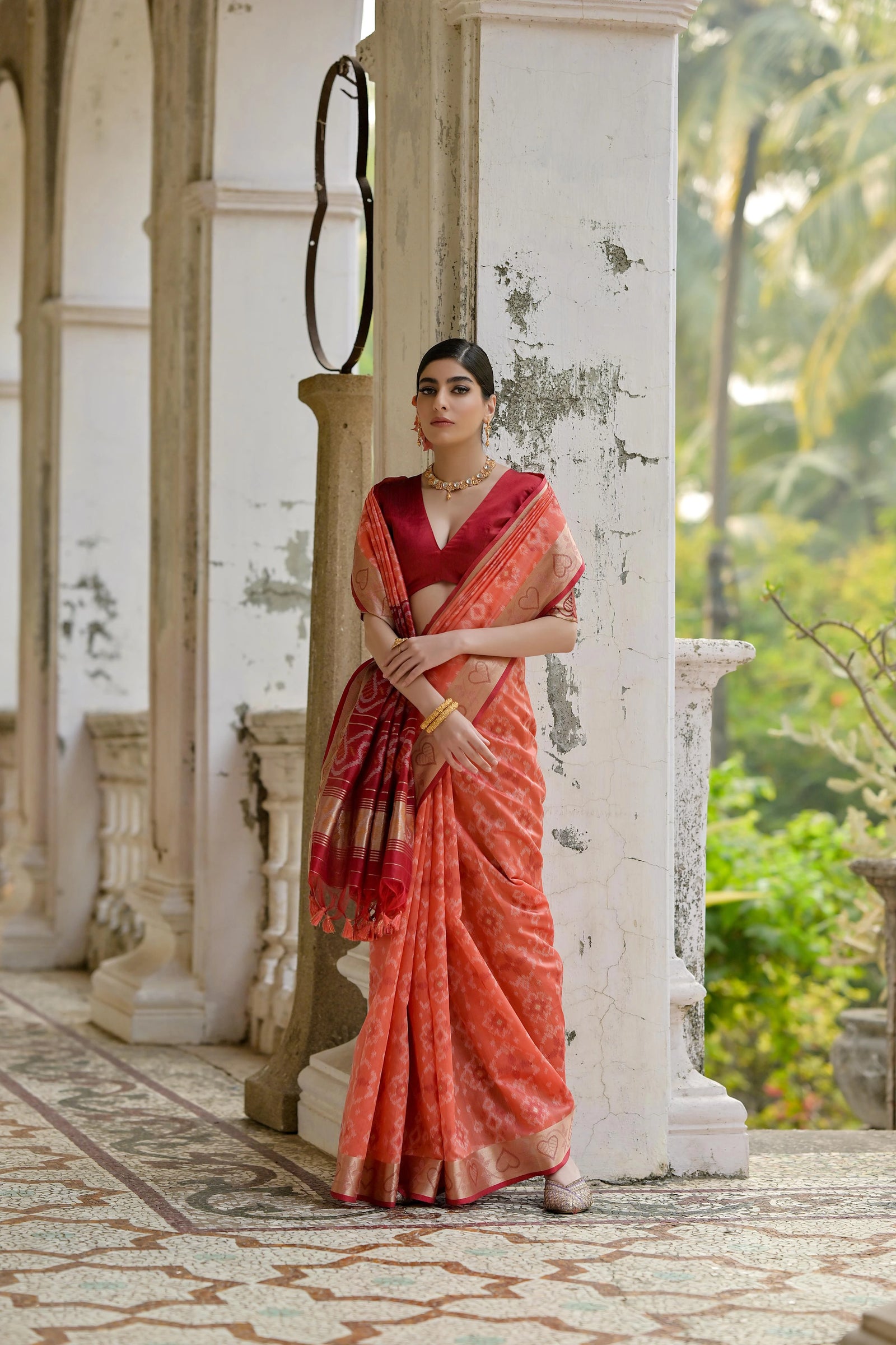 Woman in a red saree standing in an outdoor setting with columns and greenery.