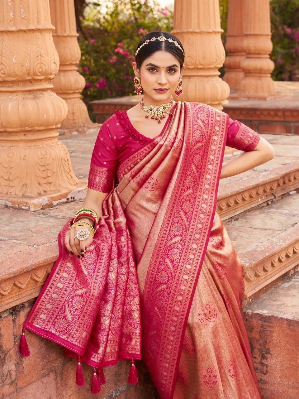 Woman in a pink saree with intricate patterns standing in front of architectural columns.
