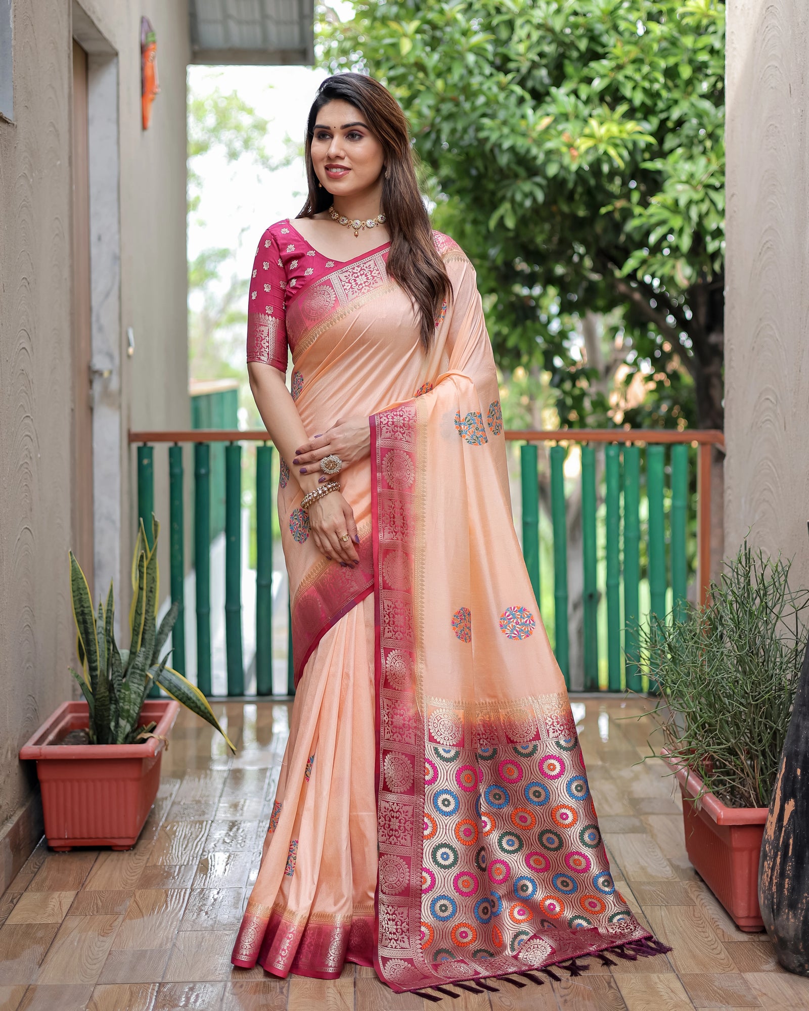 Woman in a peach and pink saree standing outdoors with plants and a building in the background