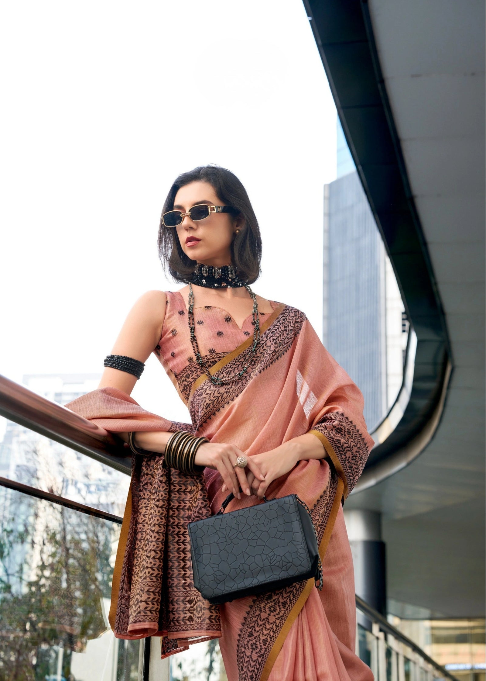 Woman in a pink saree with a black clutch standing in an urban setting.