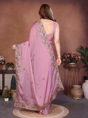 Woman wearing a pink saree with floral patterns in a room with decorative plants and vases.