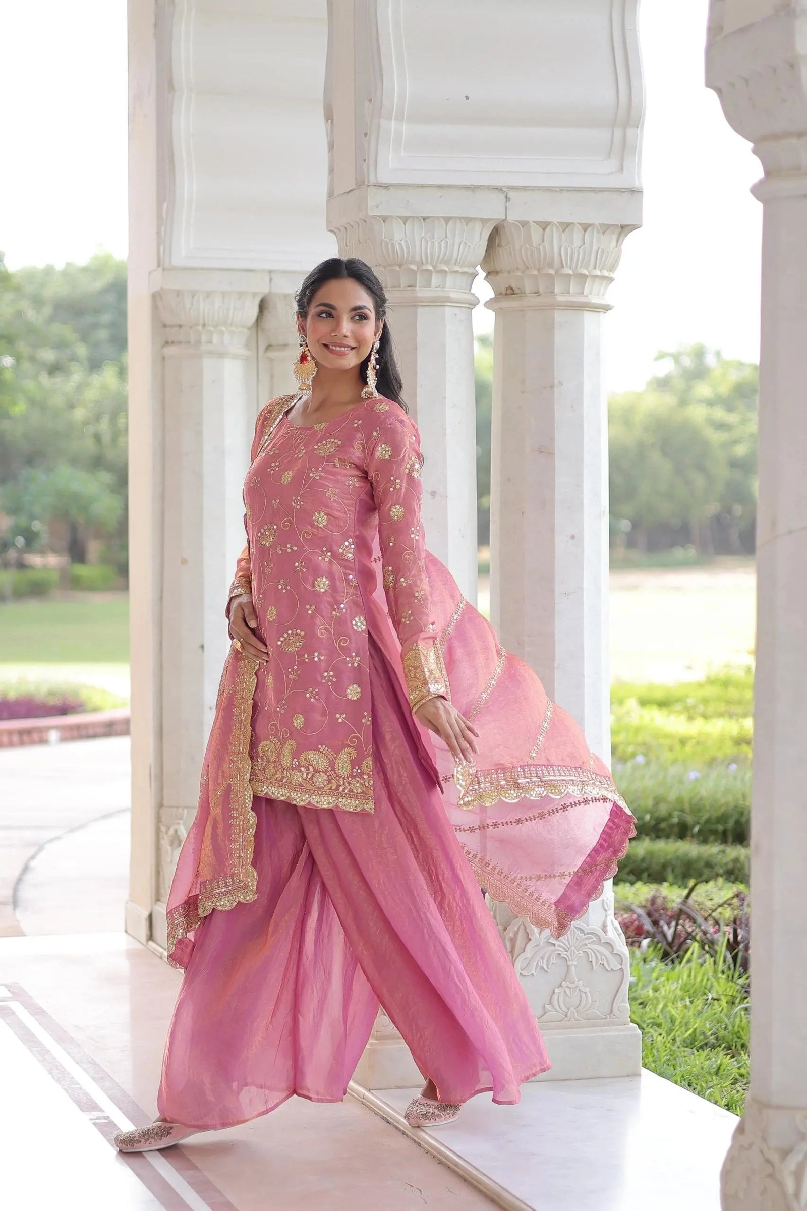 Woman in a pink traditional outfit standing in front of an architectural structure with greenery in the background