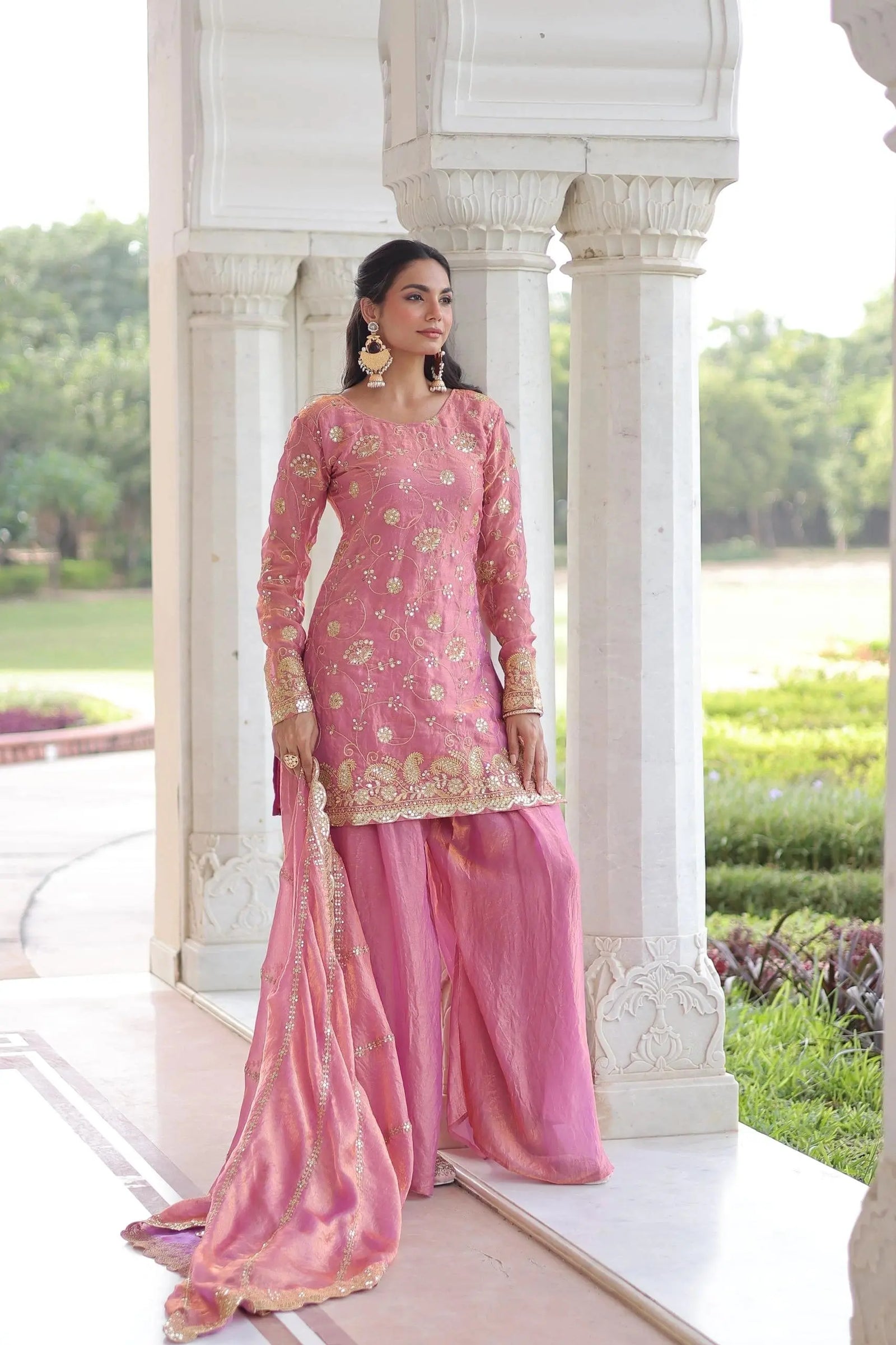 Woman in a pink traditional outfit standing in front of white architectural columns with greenery in the background.