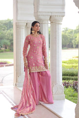Woman in a pink traditional outfit standing in front of white architectural columns with greenery in the background.