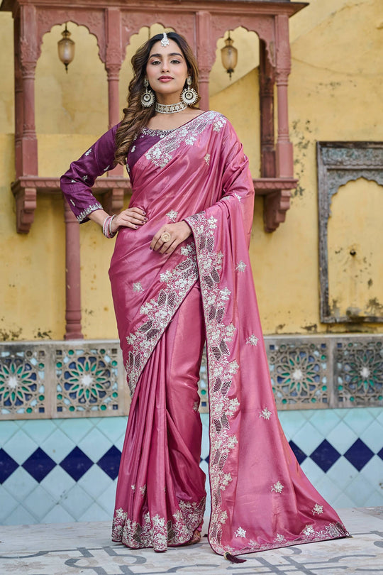 Woman in a pink saree with intricate designs standing in front of a decorative wall.