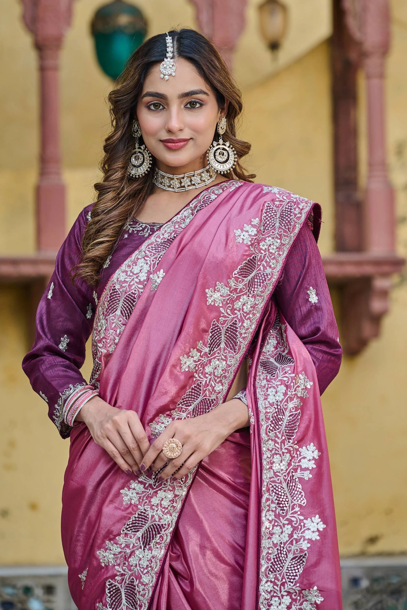 Woman wearing a pink saree with intricate designs and jewelry in an indoor setting.
