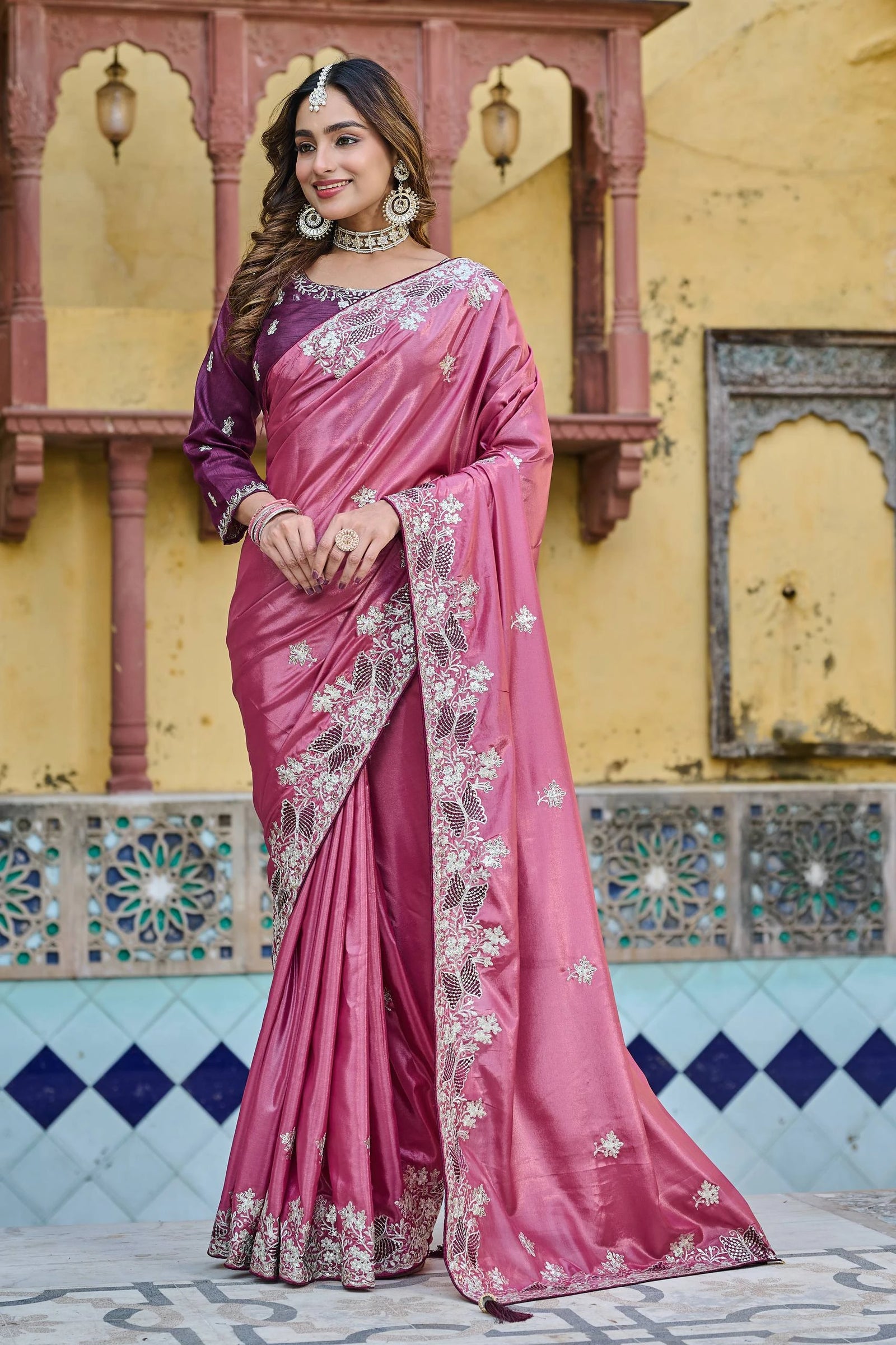 Woman wearing a pink saree with intricate designs in front of a decorative wall.